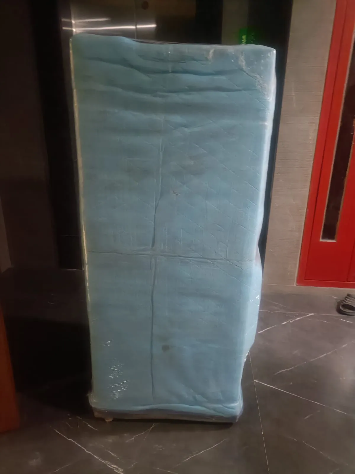 Refrigerator wrapped in a quilted blue moving blanket and stretch film beside a lift lobby